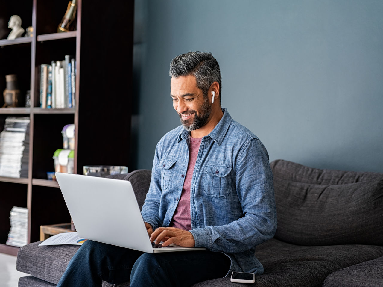 Successful mature indian businessman sitting on couch typing on laptop with wireless earphones. Mixed race businessman sitting on couch while working from home during video call. Happy multiethnic business man in casual clothing working on computer. 